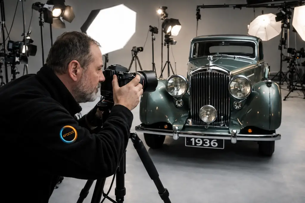 Photographer with a DSLR on a tripod shoots a vintage green car in a studio with bright studio lights.