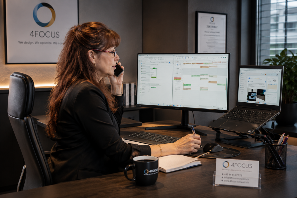 Professional woman in a black blazer sits at a desk with two large monitors, talking on the phone while taking notes. 4FOCUS branding is visible on the wall behind her.