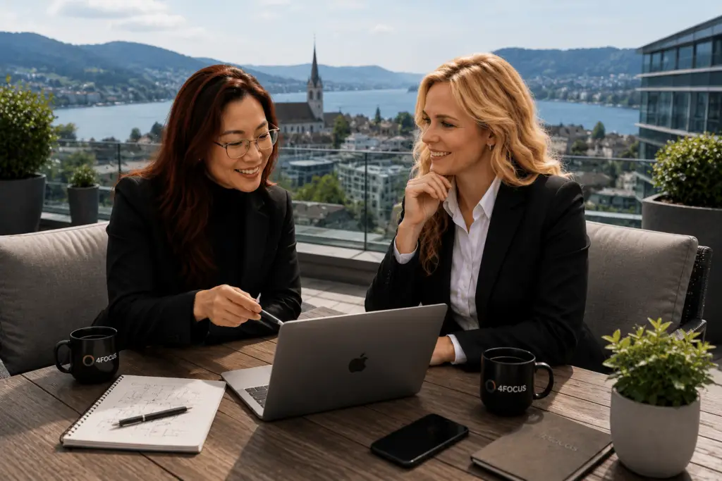 Two women in blazers sit at a wooden table on a rooftop terrace, discussing with a laptop and notebook, overlooking a lake and town.